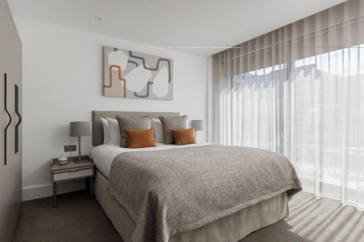 A hotel room with a large bed, a bedside table with a lamp, a wall-mounted abstract art piece, and a window with sheer curtains.