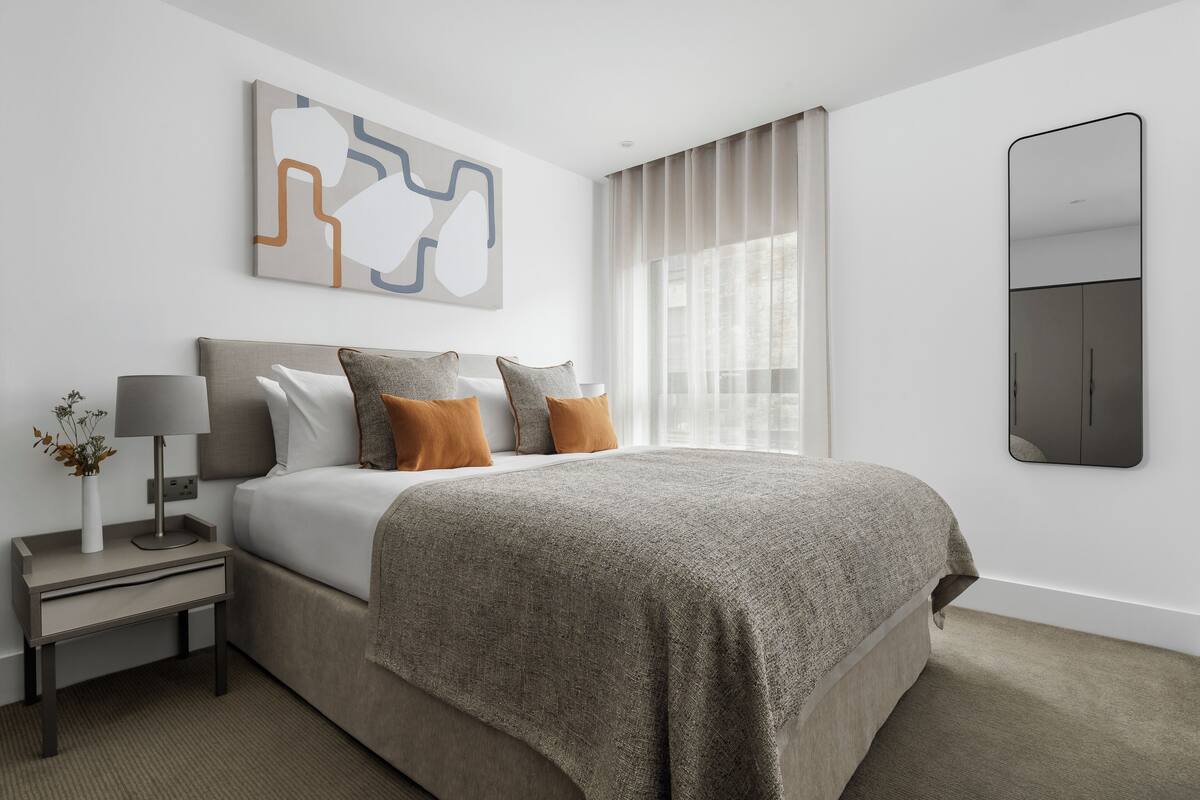 A modern bedroom with a large bed, a bedside table with a lamp, and a wall-mounted mirror.