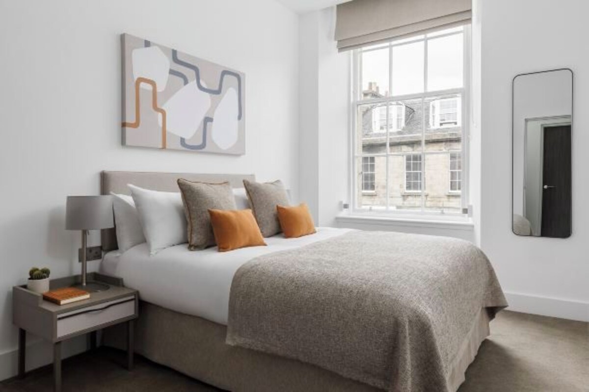 A hotel room with a bed, a nightstand, a lamp, a window with a view, and a wall-mounted mirror.