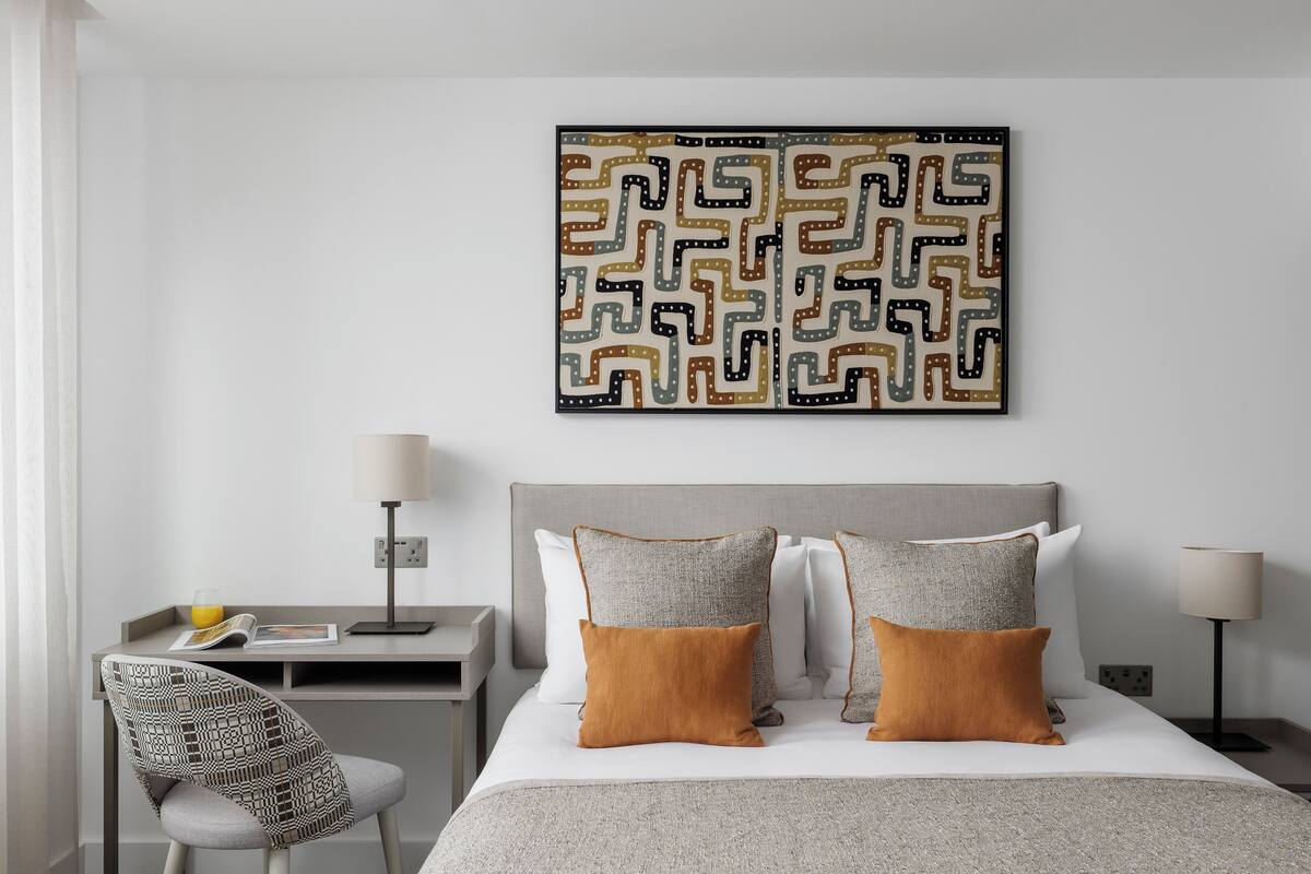A neatly arranged hotel room with a bed, two pillows, a nightstand, a lamp, a chair, and a wall-mounted artwork.