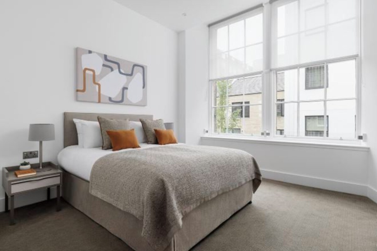 A bedroom with a large bed, a nightstand, a lamp, and a window with a view of buildings.