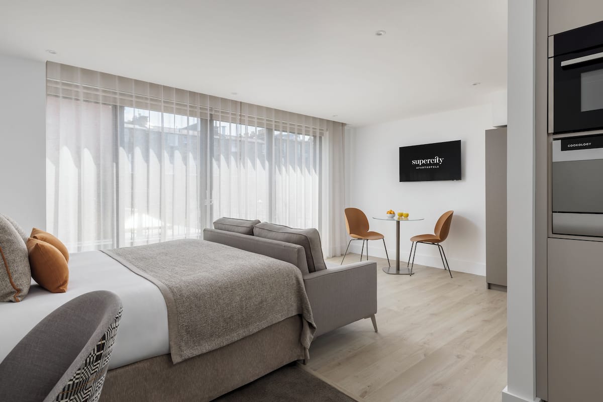 A modern hotel room with a large bed, a small sofa, a dining area with two chairs, and a TV on the wall.