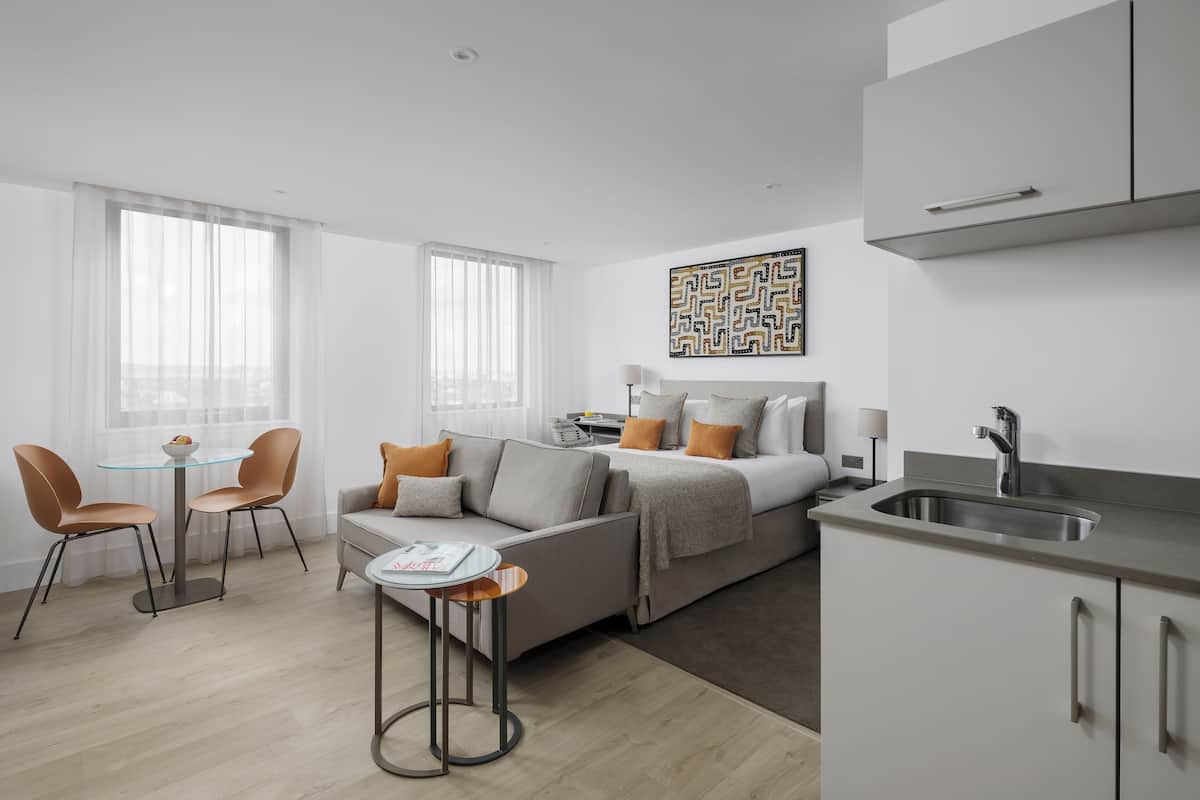 A modern hotel room with a bed, sofa, dining table, and kitchenette.