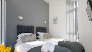 A hotel room with two beds, grey bedding, white pillows, and a grey headboard. There are two wall-mounted light fixtures and a framed picture on the wall.