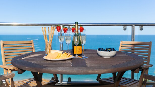A round wooden table with a bottle of champagne, glasses, a plate of cheese, crackers, and a bowl of blueberries on a balcony overlooking the ocean.