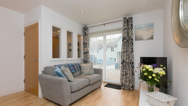 A living room with a grey sofa, a wooden door, a white wall, a window with curtains, and a vase with flowers.