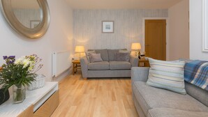 A living room with a grey sofa, a white side table with a vase of flowers, a wooden floor, and a large mirror on the wall.