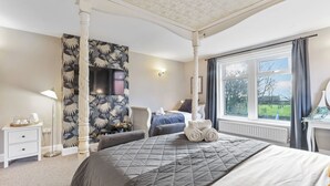 A bedroom with a four-poster bed, a flat-screen TV mounted on the wall, a large window with a view of a garden, and a bedside table with a lamp. - Wyndgrove House - North East Escapes (Seahouses)