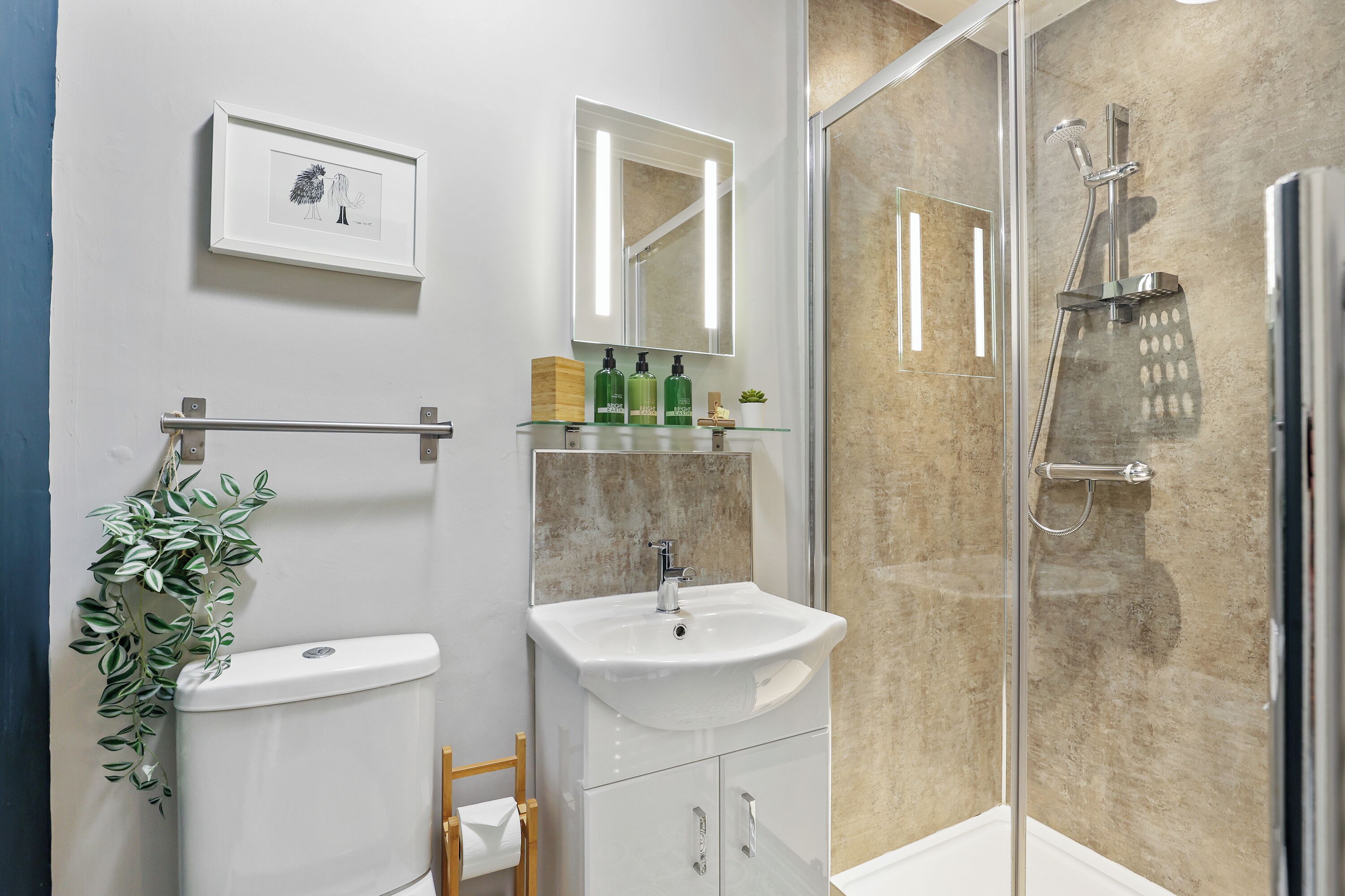 Un bagno moderno con servizi igienici, lavandino e doccia. C'è un quadro appeso al muro, una piccola pianta e articoli da toilette sul lavandino.
