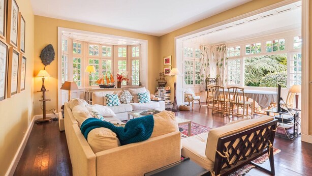 A spacious living room with a large sofa, a dining area with wooden chairs, and large windows offering a view of greenery.