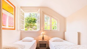 A bedroom with two single beds, a nightstand with a lamp, and a window with a view of greenery.