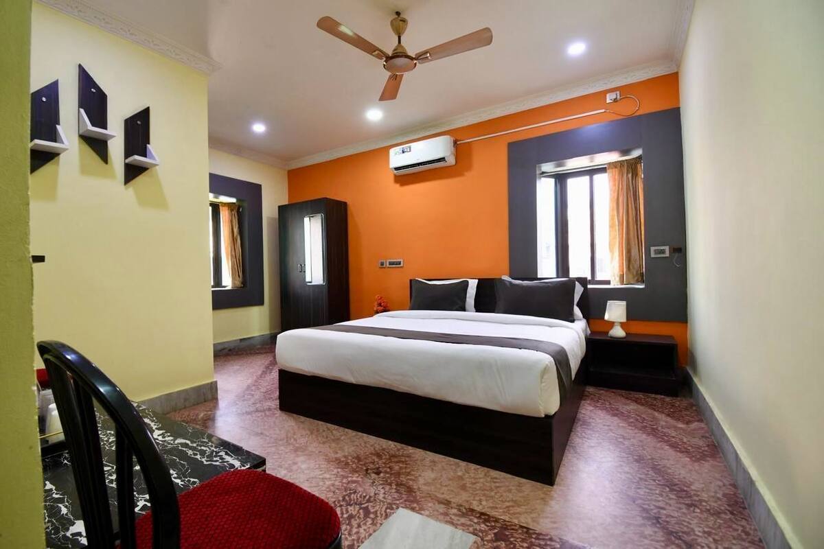 A hotel room with a bed, a ceiling fan, an air conditioning unit, a window with curtains, and a red chair.