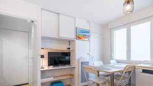 Une cuisine compacte dotée d’un téléviseur intégré, un coin repas avec une petite table et des chaises, et une fenêtre laissant entrer la lumière naturelle.