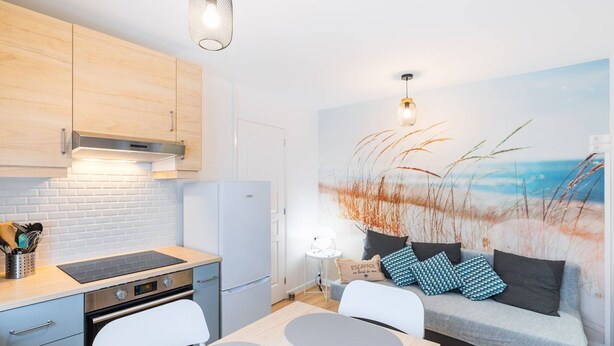 Une cuisine compacte dotée d’appareils électroménagers modernes, un coin repas avec une table et des chaises, et un espace de vie avec un canapé et une fresque murale sur le thème de la plage.