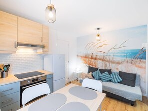 Une cuisine compacte dotée d’appareils électroménagers modernes, un coin repas avec une table et des chaises, et un espace de vie avec un canapé et une fresque murale sur le thème de la plage.