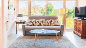Ein Wohnzimmer mit einer Couch, einem Couchtisch, einem Fernseher und Blick ins Grüne.