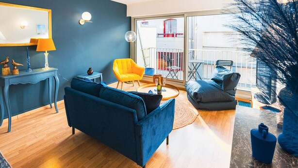 A modern living room with a blue sofa, a yellow armchair, and a dining table. There is a balcony with a view of a building. - Apartment with sea side view, city center (Le Touquet-Paris-Plage)