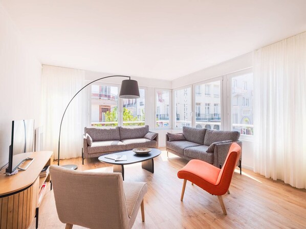 Ein modernes Wohnzimmer mit zwei Sofas, einem Couchtisch und einem Fernseher auf einem Holztisch. Große Fenster bieten Blick auf die Stadt.