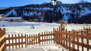 A wooden deck with a snow-covered table and benches, overlooking a snowy landscape with buildings and a forested mountain in the background. - Cozy studio at the foot of the slopes with terrace (Isola 2000)