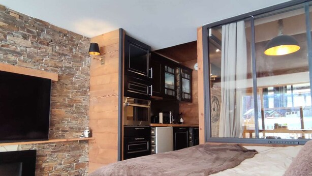 A bedroom with a large bed, a TV mounted on a stone wall, a kitchen area with cabinets, and a large window with a view. - Cozy studio at the foot of the slopes with terrace (Isola 2000)