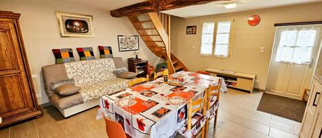 A cozy living room with a sofa, a dining table with chairs, a staircase, and a wooden ceiling beam.