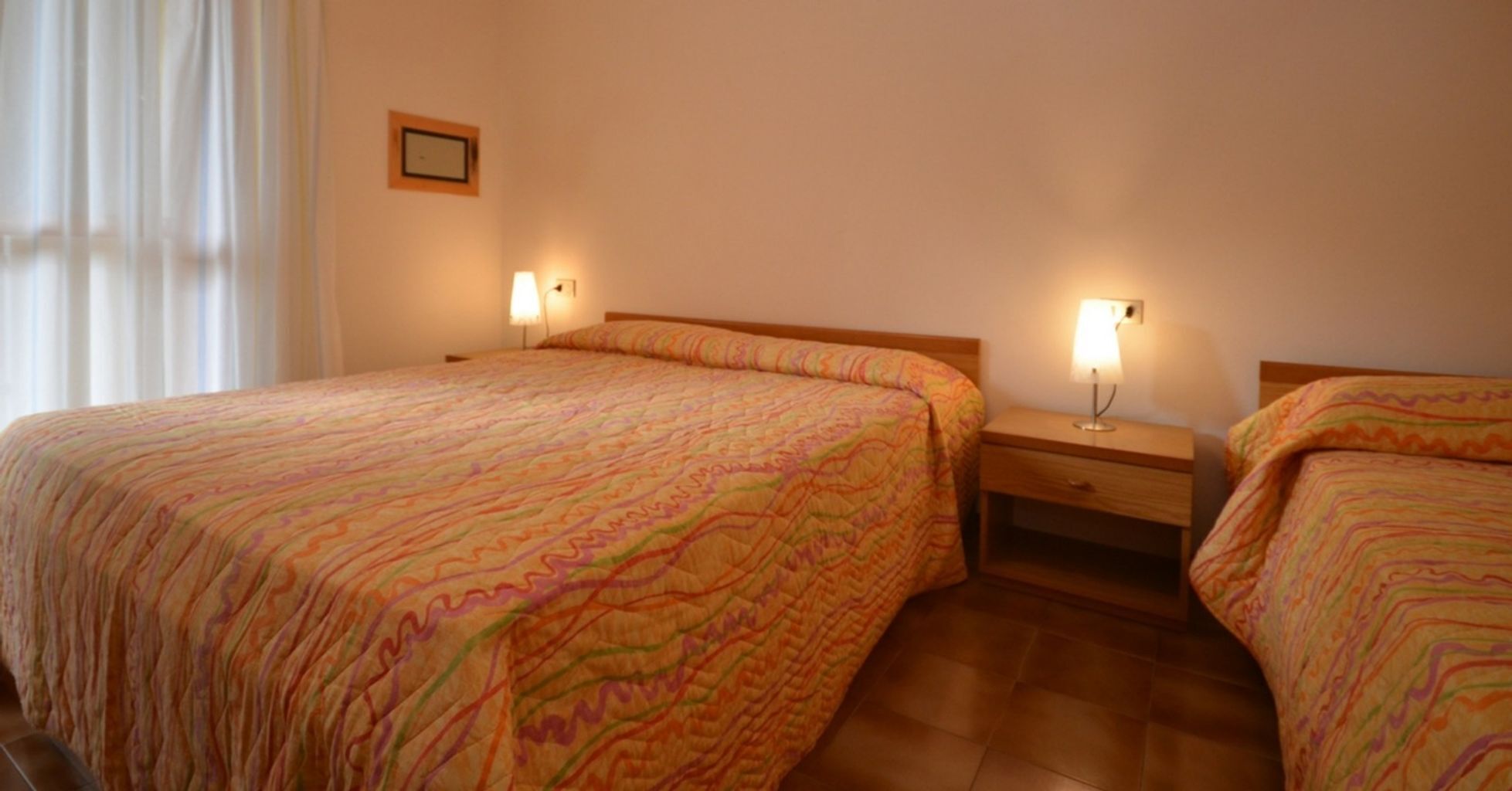 A hotel room with two beds, each with a patterned bedspread. There are bedside tables with lamps and a small framed picture on the wall.