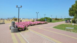 A paved pathway with a bench and pink flowers, leading to a beach area with a playground and a bicycle parked nearby.