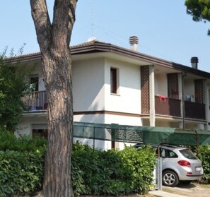 Un edificio residencial de dos pisos con un balcón, un coche estacionado al frente y un árbol a la izquierda.
