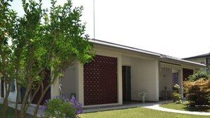 A modern building with a covered patio area, a small garden with a tree and purple flowers, and a clear sky.