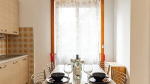 A dining area with a table set for two, a bottle of wine, and a window with a floral curtain. - Moncler House Navigli (Milano)