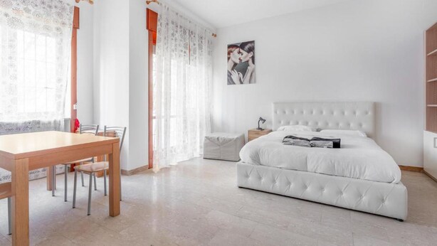 A bedroom with a white bed, a wooden table with chairs, and a window with sheer curtains. - Moncler House Navigli (Milano)