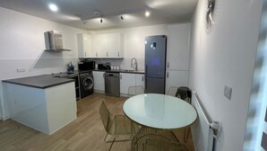 Una cocina moderna con armarios blancos, horno empotrado, refrigerador y lavavajillas. Hay una mesa redonda con sillas en el centro.