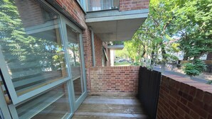 Un estrecho pasillo exterior con piso de madera, paredes de ladrillo y una puerta de cristal que da a un balcón.