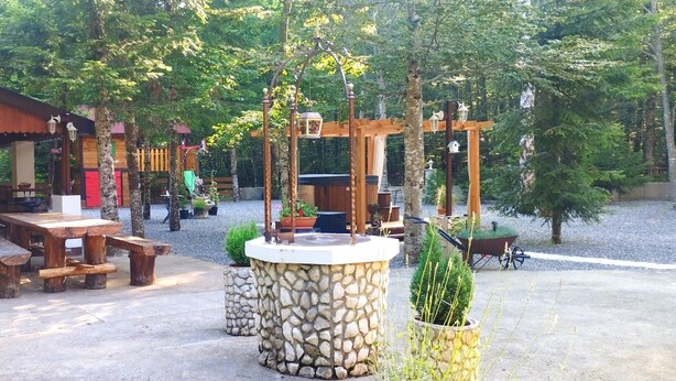 Un área de estar al aire libre con bancos y mesas de madera, una fuente de piedra y una pérgola de madera con faroles colgantes.