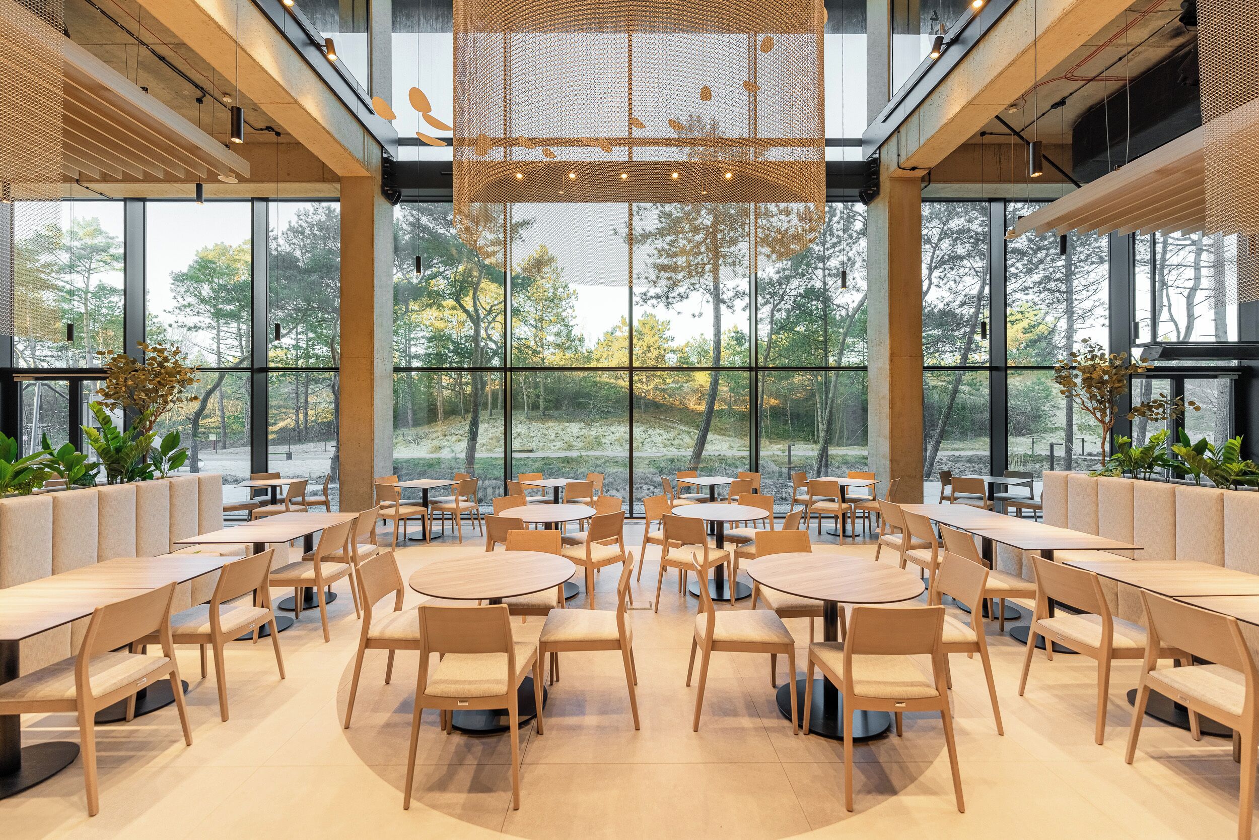 A modern indoor space with round tables and chairs, large windows offering views of trees, and a ceiling with a woven light fixture.