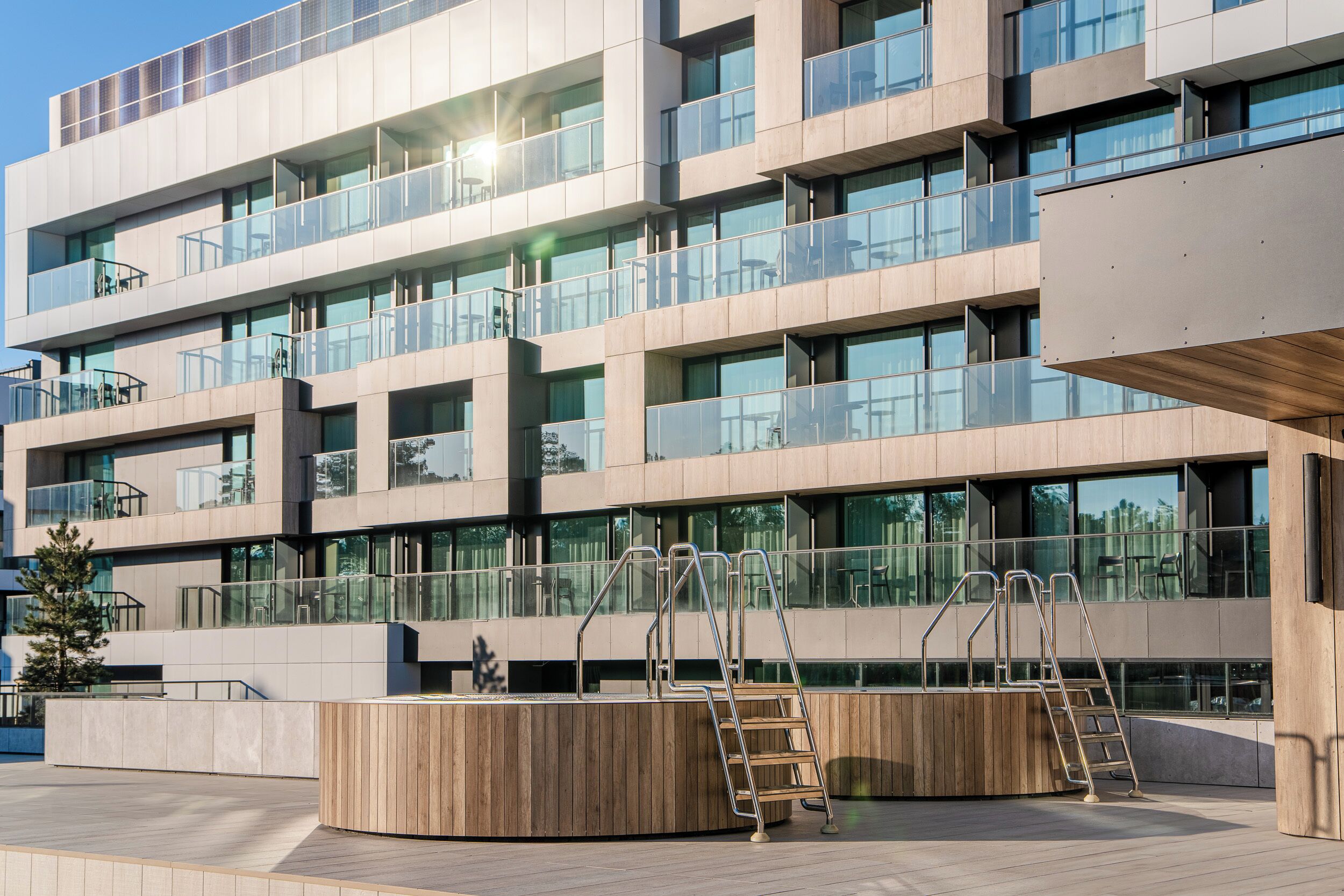 Un edificio moderno de varios pisos con balcones, ventanas amplias y una zona de terraza de madera con barandillas metálicas.