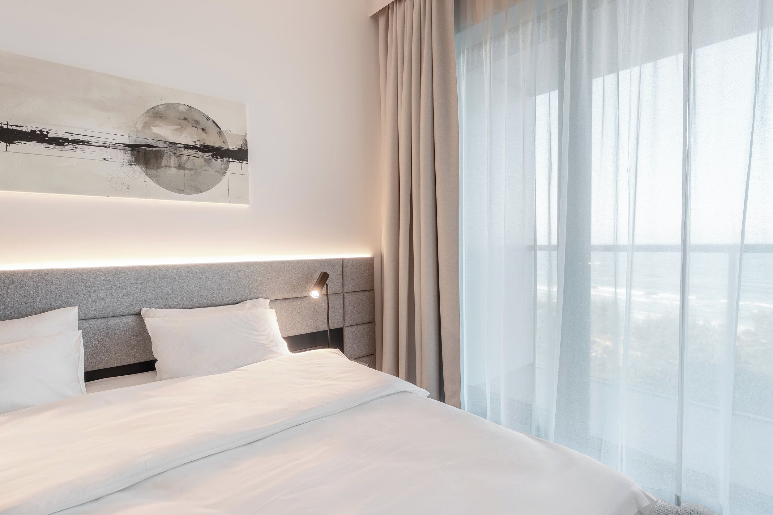 Une chambre d’hôtel moderne dotée d’un grand lit, d’une tête de lit avec éclairage intégré, d’une grande fenêtre avec des rideaux légers et d’une œuvre d’art abstraite accrochée au mur.
