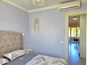 A bedroom with a tufted headboard, a bed with patterned bedding, a wall-mounted dreamcatcher, and an air conditioning unit.