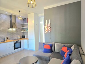 A modern kitchen with a microwave, kettle, and built-in oven. There is a grey sofa with orange and blue cushions, a small round table, and a wall-mounted light fixture.