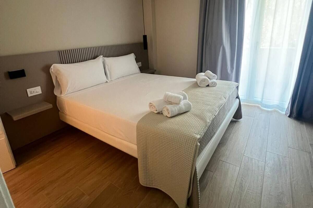 Hotelkamer met een groot bed, nachtkastje, raam met gordijnen en een ventilatie rooster in het plafond.