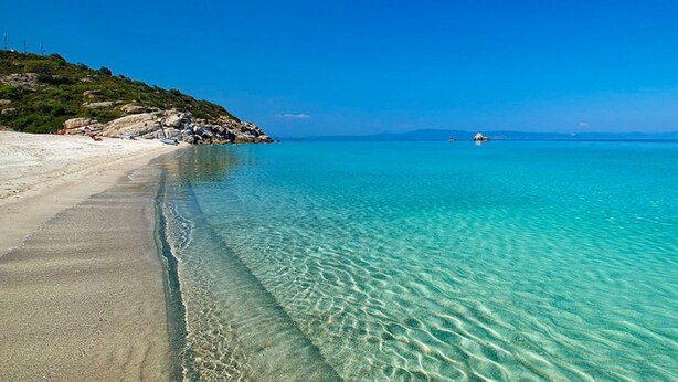 Une plage à l’eau turquoise et limpide, avec un rivage sablonneux et un terrain rocheux.