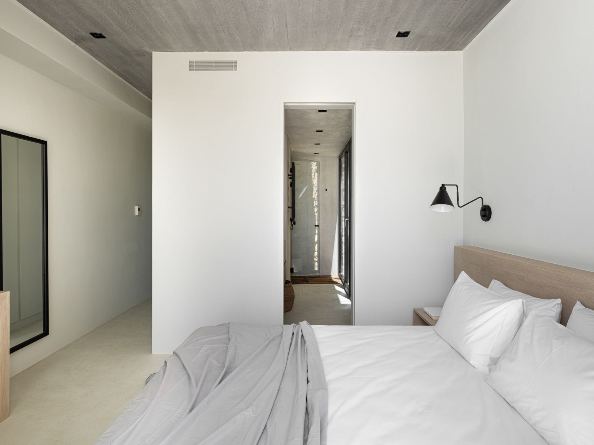 A modern hotel room with a large bed, a bedside table, a nightstand, a wall-mounted lamp, and a view of a hallway.