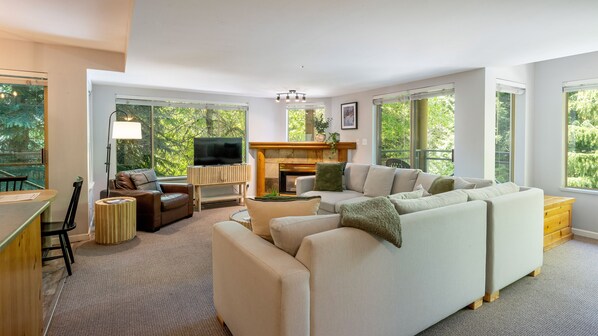 A modern living room with a fireplace, a television, and a large window offering a view of greenery.