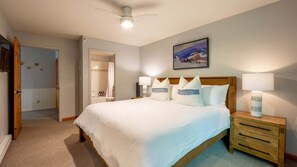 A hotel room with a large bed, bedside lamps, a ceiling fan, a painting on the wall, and a bathroom visible through an open door.