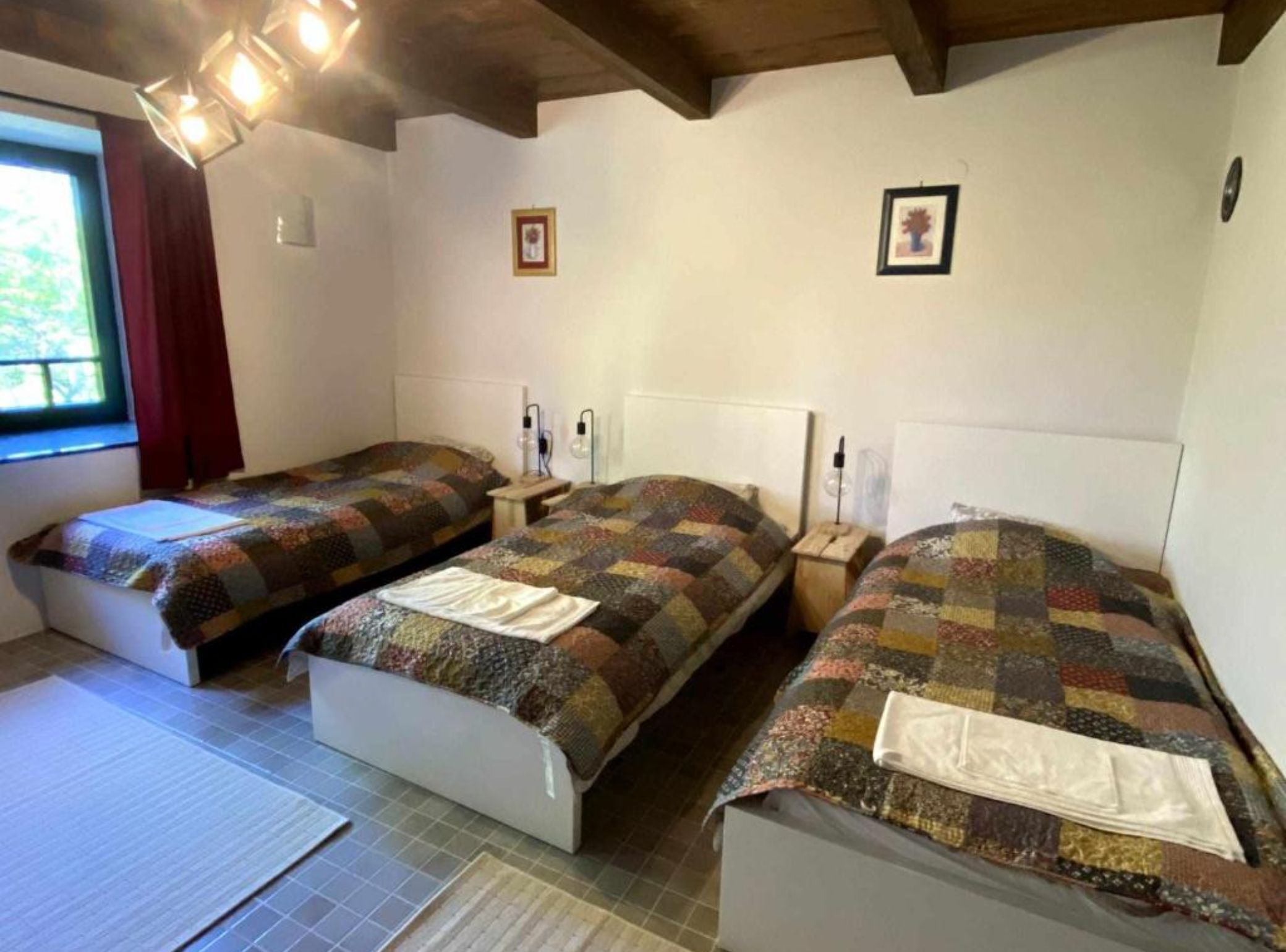 A room with three beds, each with a quilted blanket, a window with red curtains, and a ceiling with exposed beams.