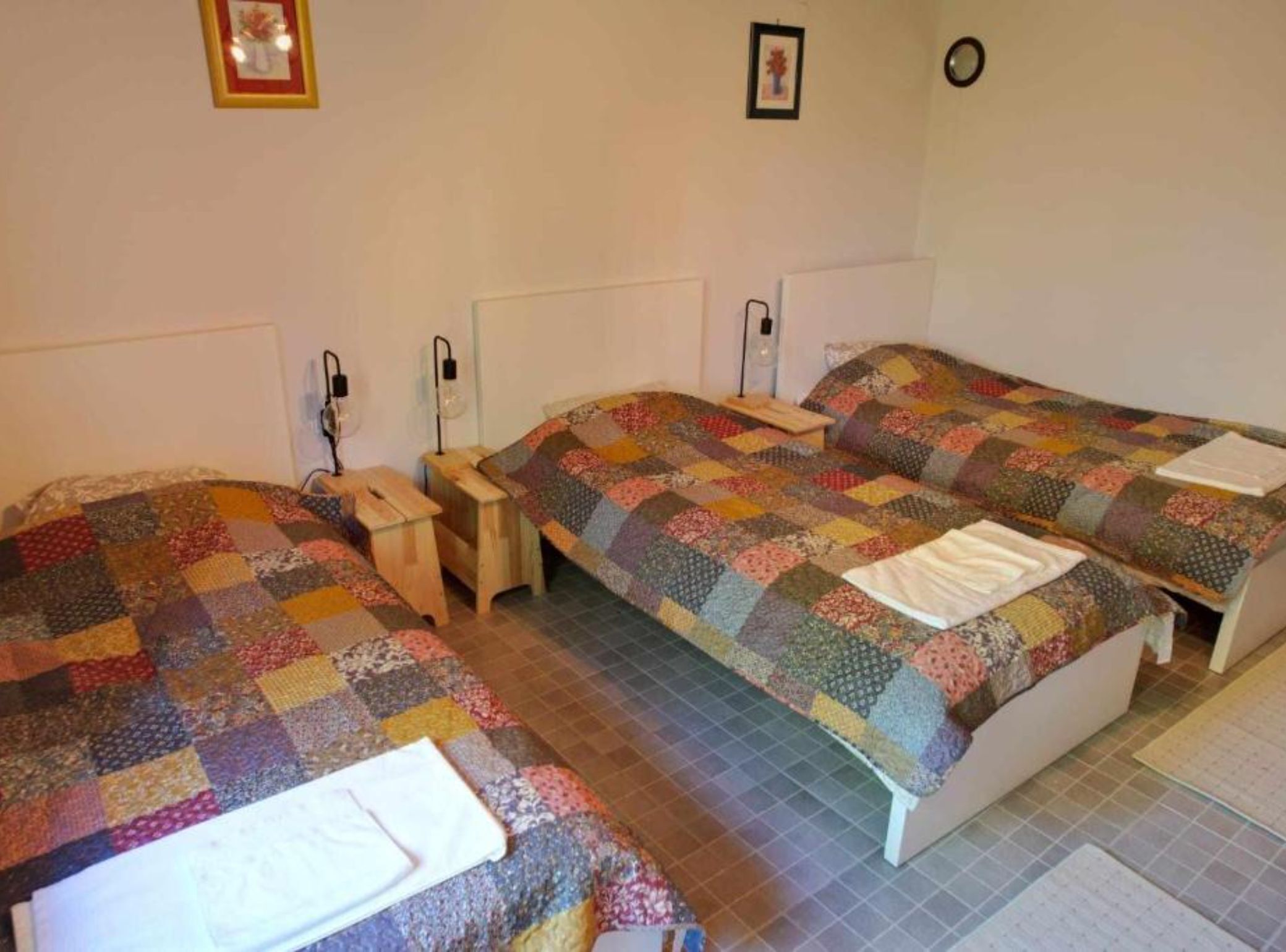 Una habitación con tres camas, cada una con un colcha de colores. Hay mesitas de noche con lámparas y un taburete de madera.