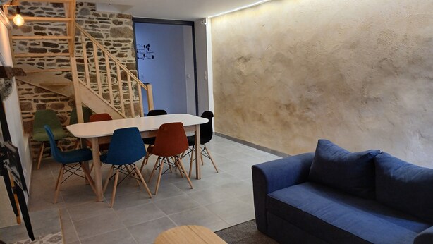 Una sala de estar acogedora con un sofá azul, una mesa de centro de madera y una zona de comedor con una mesa blanca y sillas coloridas. Una escalera con barandilla de madera conduce a una puerta con marco azul.