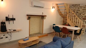Una sala de estar moderna con un sofá azul, una mesa de centro de madera, una chimenea, una mesa de comedor con sillas coloridas, un televisor montado en la pared y una escalera de madera.