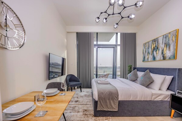 A modern hotel room with a large bed, a dining table with plates and a wine glass, a chair, a TV, and a view of the city through large windows.