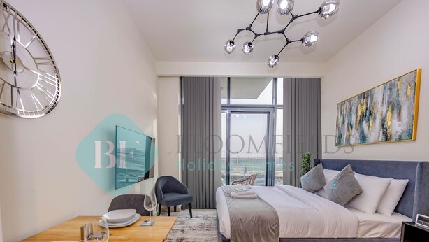 A modern hotel room with a large bed, a dining table with plates and wine glasses, a clock, and a chandelier. - Bloomfields Fantastic STU Azizi Riviera (Dubai)